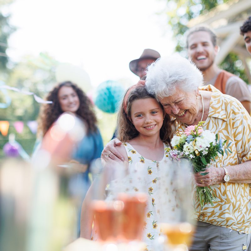 Garden birthday party for senior lady. Beautiful senior birthday woman receiving flowers from granddaughter.