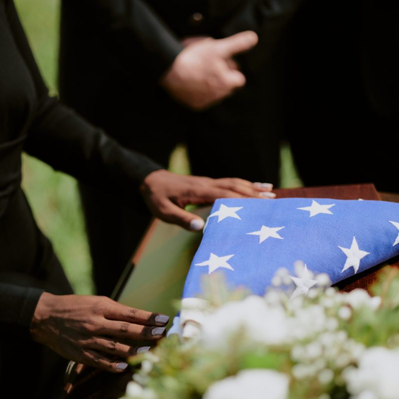 Black woman placing hands on casket draped with folded American flag during funeral ceremony, two Caucasian men standing nearby with hands clasped, floral arrangement in foreground
